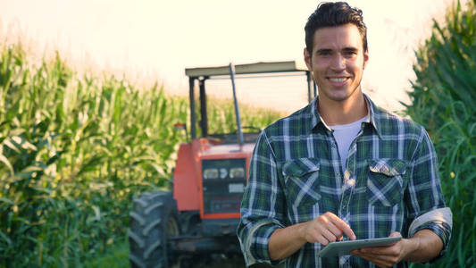 一位美丽的年轻农民学生在田里工作的肖像，拖拉机在平板电脑里工作，在衬衫玉米地里快乐。 概念生态运输农民清洁空气食品生物制品