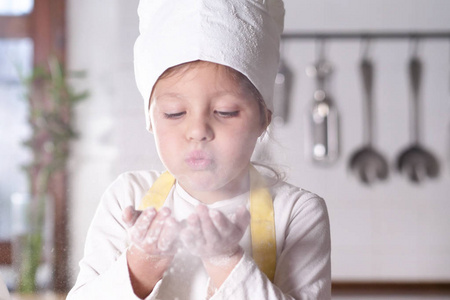 厨房里一个小女孩的肖像打扮成职业厨师，玩面粉，她的手弄脏了，笑得很开心。 概念营养烹饪学校教育与游戏。