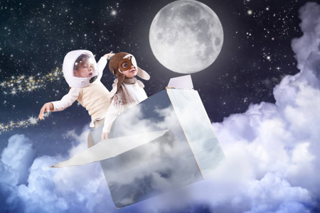 卧室里的两个小女孩打扮成飞行员或飞行员，假装驾驶纸飞机飞过云层，触摸星星，想象她在天空中自由飞翔。