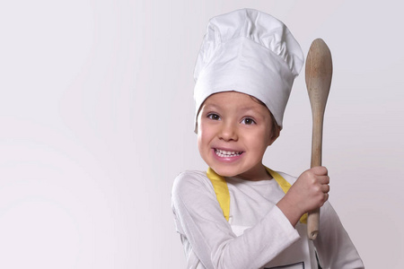厨房里一个小女孩的肖像，打扮成专业厨师，微笑着，手里拿着一个传统的木制厨房勺子。 概念营养烹饪学校教育和家庭。