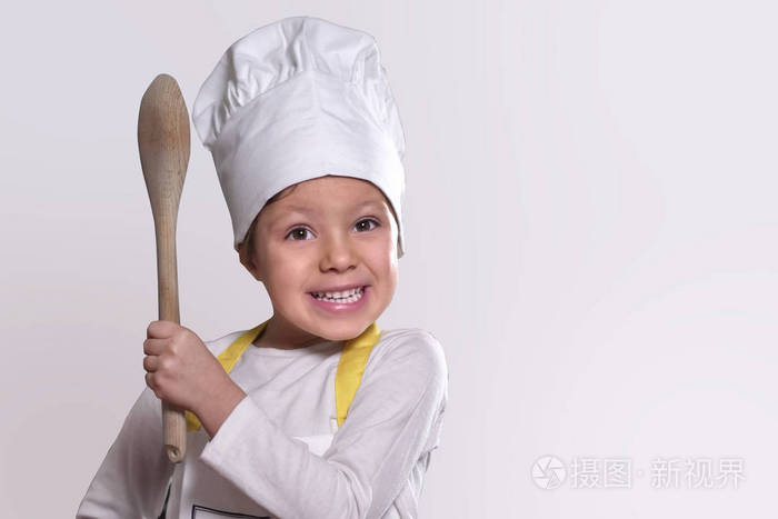 厨房里一个小女孩的肖像，打扮成专业厨师，微笑着，手里拿着一个传统的木制厨房勺子。 概念营养烹饪学校教育和家庭。
