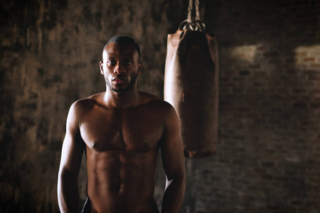 英俊赤膊肌肉发达的非洲拳手男子在镜头前摆姿势，站在拳击袋前