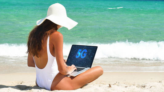 美丽的女人坐在沙滩上，在电脑上工作，带着互联网5G，戴着一顶白色的帽子，穿着白色的泳衣，背景是深蓝色的海水。