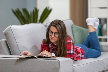 戴眼镜的女孩在家里的客厅里用耳机听智能手机上的音乐。 读杂志书。
