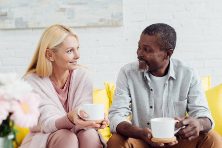 微笑的成熟女人和非裔美国人坐在沙发上，一起喝咖啡