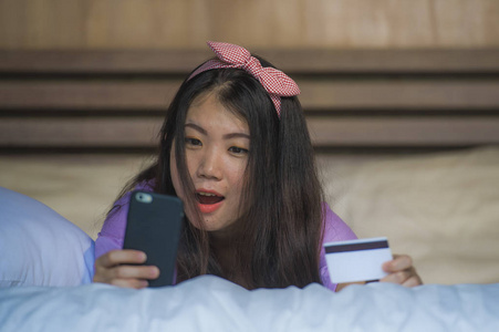 年轻美丽快乐的亚裔韩国女性在家卧室里使用信用卡网上银行和手机，在床上微笑，在电子商务商业和网上购物中快乐