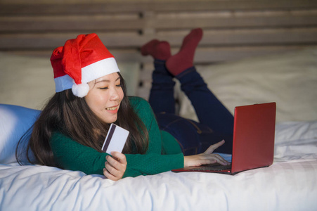 年轻快乐美丽的亚洲女孩戴着圣诞帽，用信用卡和笔记本电脑上网购物，圣诞礼物和礼物坐在床上，快乐而兴奋