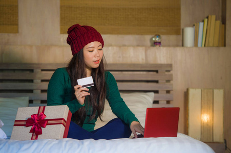 年轻美丽快乐的亚裔美国妇女在家中床上拿着信用卡和圣诞礼品盒网上购物圣诞礼物，微笑着满足电子商务和电子商务的概念