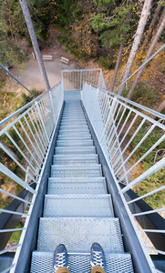 从一个观景台上看到通往森林的金属楼梯的垂直景色，前景是男脚穿蓝色鞋子