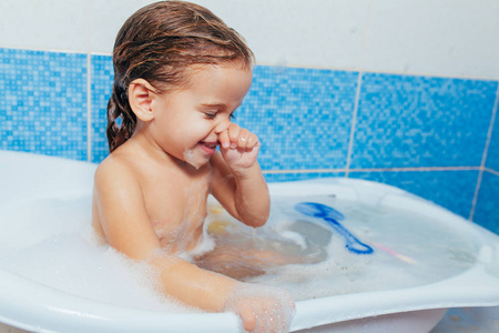 有趣的开朗快乐幼儿宝宝洗澡时玩泡沫泡泡。浴缸里的小孩微笑的孩子在浴室在蓝色背景。卫生和保健