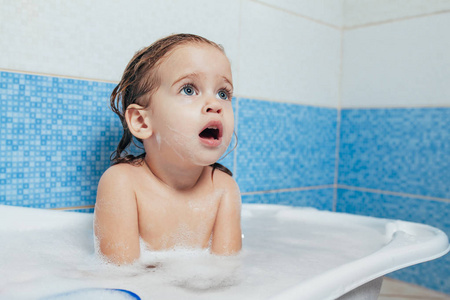 有趣的开朗快乐幼儿宝宝洗澡时玩泡沫泡泡。浴缸里的小孩微笑的孩子在浴室在蓝色背景。卫生和保健