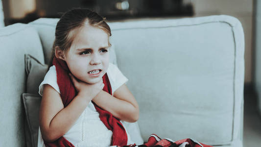 戴着红领巾坐在沙发上的小女孩。 生病的年轻女孩。 房间里有白色沙发。 不幸的孩子。 疾病概念。 医疗保健和健康的生活方式概念。 图片