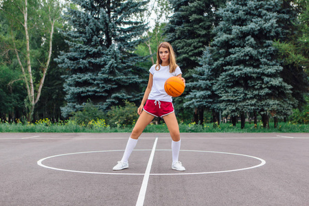 美丽的金发女孩穿着白色T恤短裤和运动鞋在户外篮球场上玩球。 复制空间。