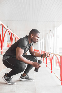 年轻的男慢跑运动员训练和做户外锻炼在城市。一个黑人在锻炼后休息, 听音乐, 看体育手表