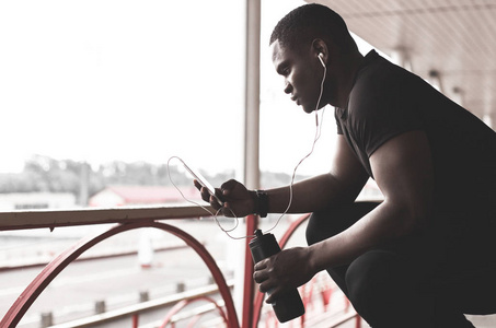 年轻的男慢跑运动员训练和做户外锻炼在城市。一个黑人在锻炼后休息, 听音乐, 看体育手表