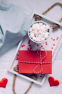 创意顶景平卧窗台生活浪漫构图。 热可可咖啡巧克力与棉花糖杯红色礼品托盘在床上。 概念情人节舒适温馨的家清晨自然光