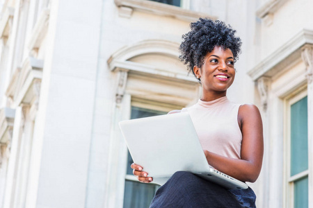 通往成功的道路。 年轻的非裔美国妇女，发型是非洲式的，穿着无袖浅色上衣，坐在纽约的老式办公楼旁，工作在笔记本电脑上，微笑着抬头