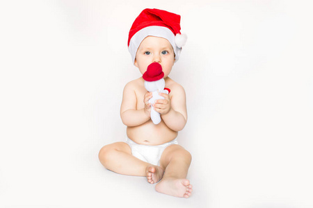 圣诞节和家庭概念，可爱，有趣的婴儿，戴着红色圣诞老人帽，坐在白色的床上，在家里幽默笑话，抱着一个小雪人