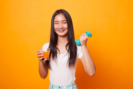 带哑铃的年轻亚洲女人喝橙汁，背景为亮黄色