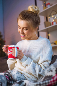 在圣诞节的时候，带着棉花糖的热可可杯子坐在沙发上，裹着毯子的年轻金发女人微笑着