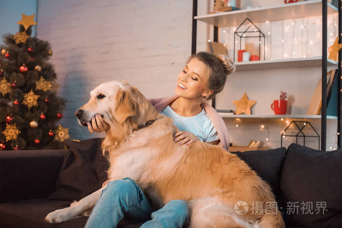 圣诞节时美丽快乐的金发女郎坐在沙发上拥抱着金毛猎犬