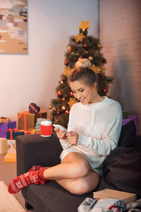 年轻的金发女郎微笑着坐在沙发上，圣诞节时使用智能手机