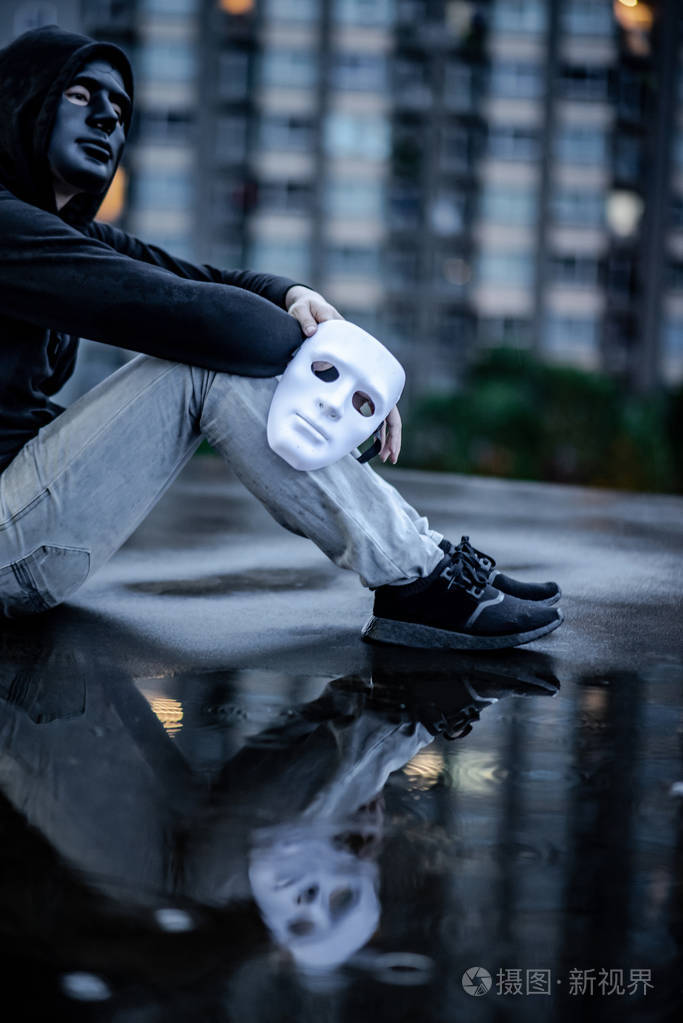 神秘的帽衫男子与黑色面具手持白色面具坐在雨中的屋顶上废弃的建筑