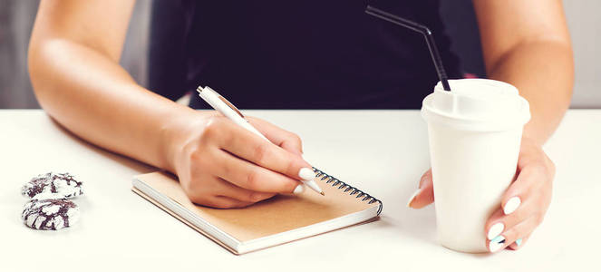 s hand writing in notepad placed on white surface with coffee cu