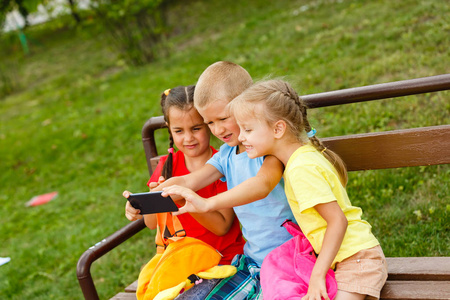 三个快乐的孩子在公园的长凳上玩智能手机