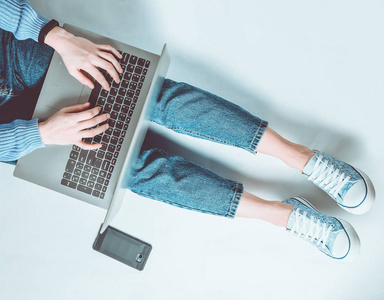在笔记本电脑上工作的博客概念。 用笔记本电脑坐在白色的地板上。 现代技术。 穿牛仔裤和运动鞋的女性腿。 上面的风景。 平躺着。