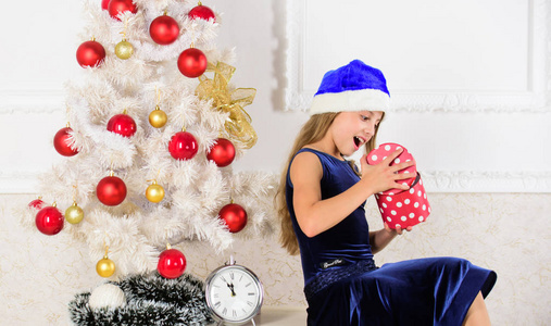 孩子女孩坐在圣诞树附近拿着礼品盒。小女孩圣诞老人帽子兴奋的圣诞礼物或礼物。是时候打开圣诞礼物了。新年快乐的概念。孩子在家里庆祝圣