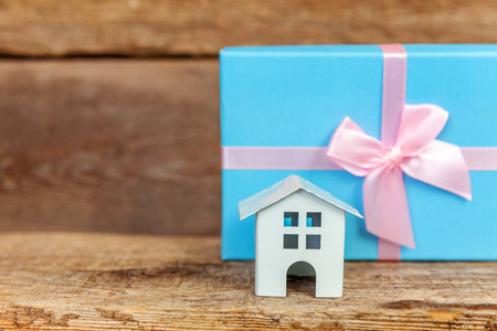 微型白色玩具屋和礼品盒包在破旧的乡村木制背景上的蓝色纸。 抵押财产保险梦想家居概念。 为家庭买新房子