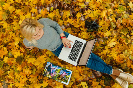 在公园里，坐在秋橙叶子上的女人用笔记本电脑和家庭相册