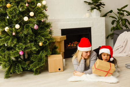 母亲和女儿戴着红色圣诞帽，礼物躺在客厅圣诞树附近的地板上