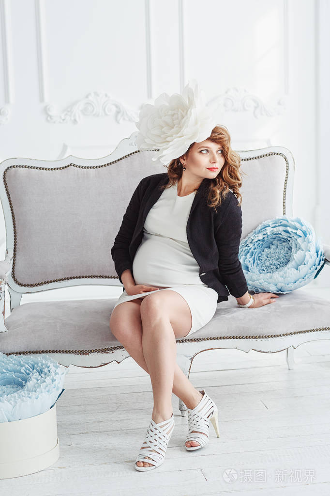 年轻优雅的孕妇的肖像她的白色大花坐在白色的沙发上抱着她的胃