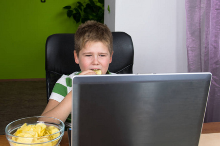 肥胖的小男孩坐在电脑前吃薯片。