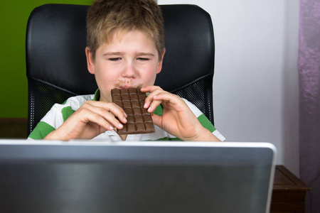 肥胖的小男孩坐在电脑前吃巧克力。