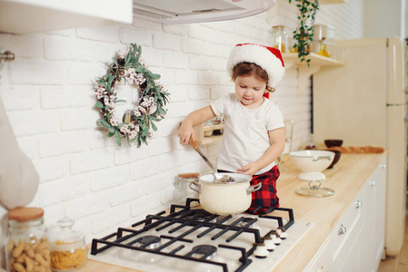 可爱的小女孩戴着圣诞帽，在家里的厨房里准备饼干。 坐在厨房的桌子上，帮妈妈准备节日的圣诞晚餐