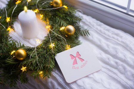 贺卡与最美好的祝愿附近的冷杉花环装饰着金色的圣诞球和蜡烛，并盘绕着发光的花环，温暖的灯光在白色针织格子。 新年家居装饰