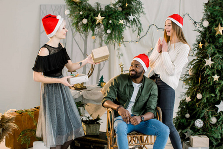 图片显示一群朋友戴着圣诞帽在家庆祝圣诞节。 穿灰色裙子的漂亮女孩给她的朋友非洲男人和高加索女人送礼物。