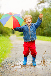 孩子穿着靴子跳上水坑。 宝贝在雨中。 一个带彩虹伞的男孩正在外面走。 秋季假期