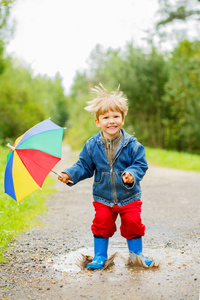 孩子穿着靴子跳上水坑。 宝贝在雨中。 一个带彩虹伞的男孩正在外面走。 秋季假期