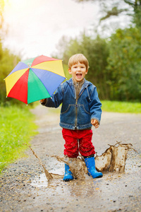 孩子穿着靴子跳上水坑。 宝贝在雨中。 一个带彩虹伞的男孩正在外面走。 秋季假期