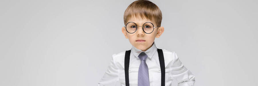 一个迷人的男孩穿着白衬衫, 吊带, 领带和浅色牛仔裤站在灰色的背景。戴眼镜的男孩