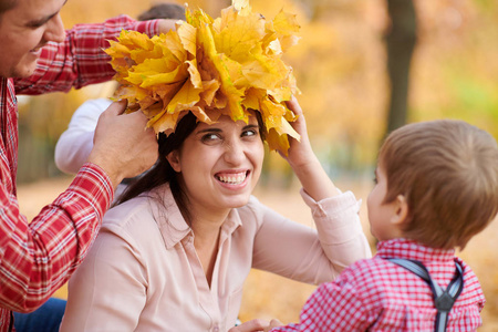 父子把黄色的落叶放在母亲的头上。 幸福的家庭在秋天的城市公园。 孩子和父母。 他们摆出微笑的姿势，玩得很开心。 明亮的黄色树木。