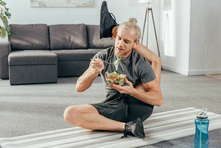 灵活的成人男子，腿后头坐在瑜伽垫上，吃蔬菜沙拉