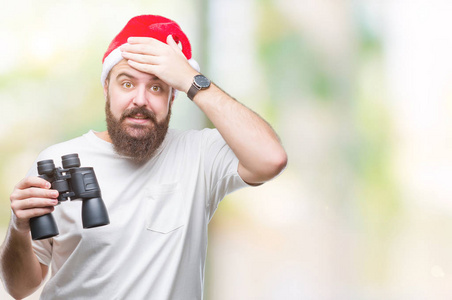 年轻的白种人嬉皮士戴着圣诞帽，看着望远镜在孤立的背景上强调，手在头上震惊，羞愧和惊讶的脸，愤怒和沮丧。 因为错误而恐惧和不安。