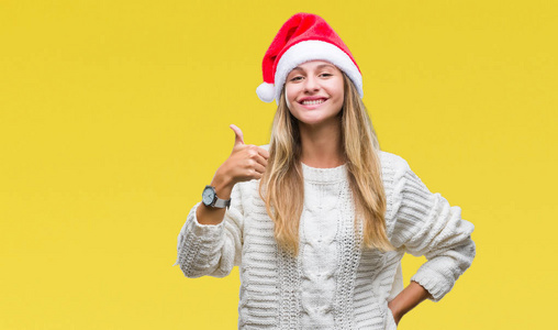年轻漂亮的金发女人戴着圣诞帽，戴着孤立的背景，用手做着快乐的竖起大拇指的手势。 赞许的表情看着相机，显示出成功。