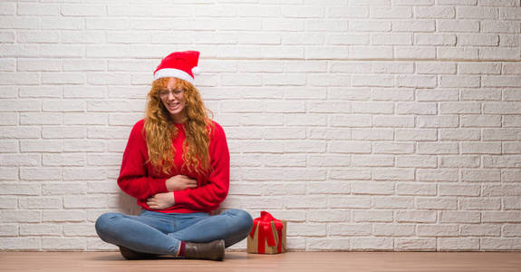 年轻的红头发妇女坐在砖墙上，戴着圣诞帽，手放在肚子上，因为消化不良，痛苦的疾病，感觉不舒服。 疼痛的概念。