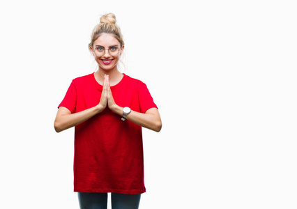 年轻漂亮的金发女人穿着红色的T恤和眼镜，在孤立的背景上祈祷，双手一起祈求宽恕，微笑自信。